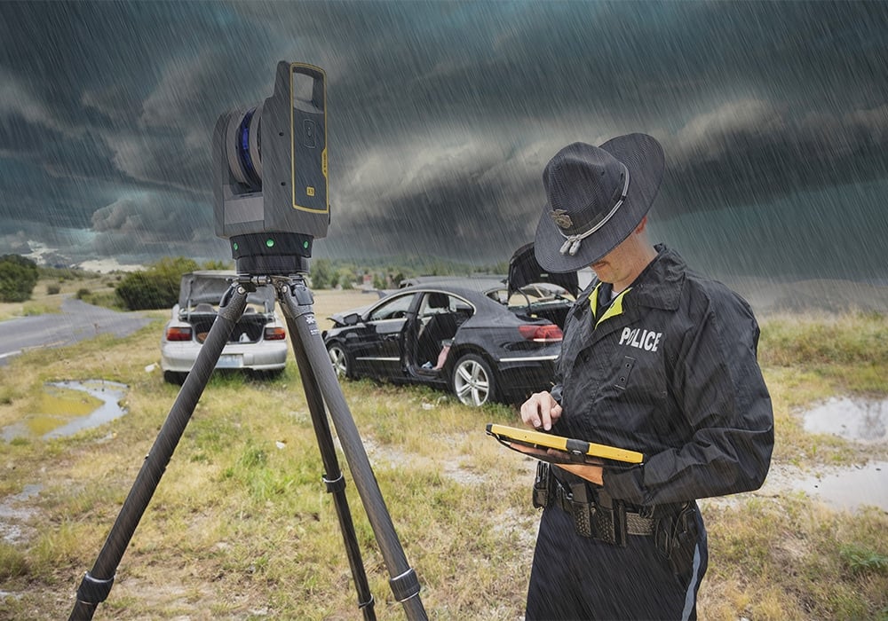 Police officer in rain using a tablet beside a tripod-mounted surveying device at a car crash scene.